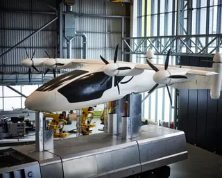 A white aircraft with multiple propellers sits on a platform inside a research hangar