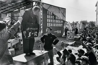 文革批鬥現場，一名長者站在桌上，頸掛一個寫有其姓名的牌子，周圍有群眾握拳。