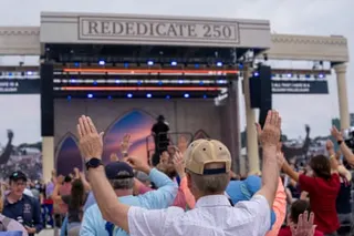 A crowd with raised hands faces a large stage with blue lights and an arched backdrop