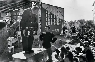 文革批鬥現場，一名長者站在桌上，頸掛一個寫有其姓名的牌子，周圍有群眾握拳。