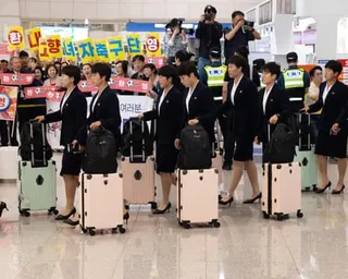 Players, all dressed in matching skirt suits and heels, wheeling suitcases through airport in front of photographers and crowds