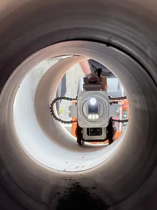 一個圓形污水管內部視角，中央是一台小型機器人，配備前置攝影鏡頭與照明燈，正在管道內行進。機器人兩側有鏈條狀部件。