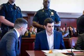 a man sits in court next to his lawyer as police stand behind him