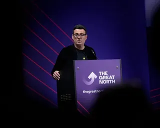 Andy Burnham speaks at an event. He stands illuminated on a dark stage with shadows around him and in front of a purple backdrop with red stripes. A sign on his lectern has an arrow logo and reads The Great North. He wears black.