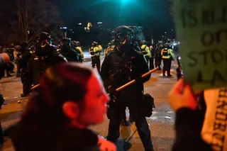 Police in riot gear with batons face protesters holding red flares on a nighttime street