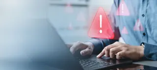A person typing on a laptop with a red warning triangle icon displayed on the screen.