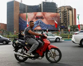 A man on a red motorbike in traffic with a large billboard and buildings in the background