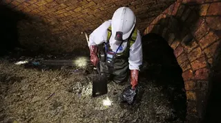 一名工人在下水道內作業，身穿全套防護裝備，包括白色安全帽、頭燈與防水工作服，站在滿佈污物的地面上。工人手持鏟工具，正挖起黑色凝固物。