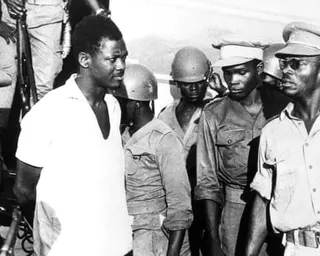 Patrice Lumumba pictured with his hands tied behind his back in 1960, surrounded by men in uniforms.