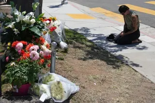 A person kneels on a sidewalk near bouquets of flowers placed beside a tree