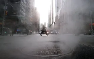 A silhouetted figure crosses a misty New York street with steam rising from a manhole cover