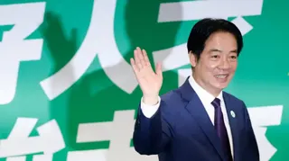 台灣總統賴清德站在台北的民進黨總部，對著鏡頭揮手微笑。他身後是帶有中文字樣的綠色背景。