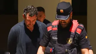Reuters Jonathan Andic wears a blue jacket and blue top while he walks with his hands behind his back and his head down. He stands next to a policeman who is wearing a black and red uniform and cap.