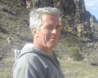 Jeffrey Epstein in a grey hoodie and red shorts stands on dirt ground with rocky cliffs behind him