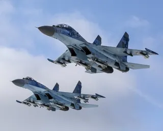 Two military fighter jets fly close together, armed with missiles, against a blue sky