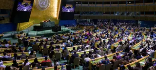联合国大会大厅全景，正值联合国原住民问题常设论坛（UNPFII）开幕之际。大厅内座无虚席，与会者坐在弧形桌前，面向悬挂着联合国大徽标的中央讲台。