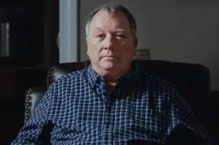 Man in a blue checked shirt sits on a dark sofa looking at the camera