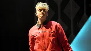 Former Why Don’t We member Jack Avery performs onstage wearing a red jacket during a live concert appearance.
