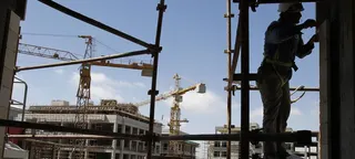 一名头戴安全帽、身系安全带的建筑工人站在建筑工地的脚手架上，背景中有起重机和正在施工的建筑。
