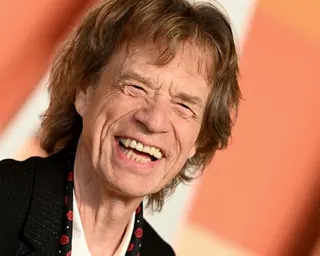 Mick Jagger laughs widely, wearing a black jacket with red decorative elements