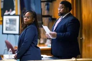 A woman in a blue suit looks off to the side as a man in a suit holds documents near her