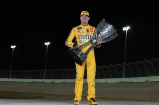 man in a yellow jumpsuit with M&M's branding smiles while holds a large trophy on a racetrack at night