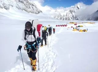 A line of people walk using poles in a snowy mountain environment. A large number of red and yellow tents can be seen in the background