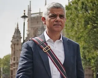 Sadiq Khan walking outdoors with a historic building and trees behind him