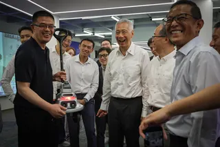 盲人工程师朱清毅（左一）举起导盲机器人，向国务资政李显龙（右三）展示它如何通过多传感器融合感知周围环境。机器人随后语音播报前方人群的衣着与外貌特征，引得现场一阵笑声。（海峡时报）