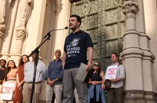 a man speaks into a microphone as people stand behind him outside a church