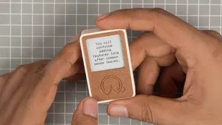 Two hands cradle a tiny epaper device. The shape of a fortune cookie is engraved into the device's 3D printed PLA housing.