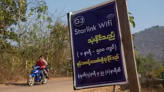 在緬甸克耶邦，一塊貼在木板上的紙質告示以英文寫著「Starlink Wifi」，並附有緬文說明。背景中可見兩人騎着一輛藍色電單車行駛在道路上，遠處則是綠樹覆蓋的山丘。（16/2/2025）