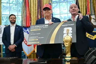 畫面攝於美國華盛頓白宮橢圓形辦公室。兩名身穿西裝的人士手持2026年世界盃決賽門票合影，其中一人戴著紅色帽子，帽上印有「Trump was right about everything」字樣。背景可見另一名人士站立在房間內，眾人位於一張辦公桌後方，而前景則擺放著世界盃獎盃