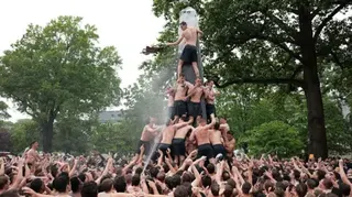 A horde of topless naval freshmen join together to climb the greased obelisk.