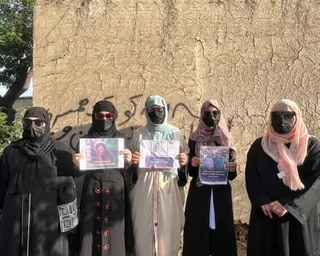 Women wearing sunglasses, face masks and hijabs stand in front of a wall holding signs