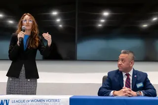 Maureen Galindo holding a microphone gestures while speaking as Johnny Garcia sits at a blue table