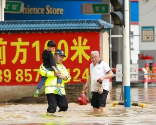 A police officer in a reflective vest carries a child through knee-deep flood water on a city street