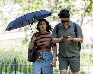 A woman holding an umbrella walks beside a man looking at his phone in a sunny park