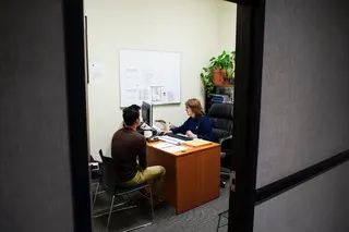 A man sits at a desk with an immigration officer in an office.
