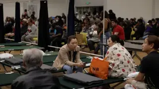 Reuters Residents sit at a temporary shelter in Fountain Valley, California. In the foreground, four people sit on dark green cots, and one woman checks her phone. Another woman is wrapped in a blanket with a pawprint and heart pattern. In the background, dozens more people sit and wait.