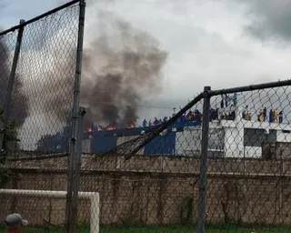Billowing smoke, fire and people on a building's roof are seen in the distance behind a broken barbed wire fence