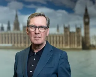 Alan Milburn in glasses and a dark suit stands outdoors with the Houses of Parliament blurred in the background