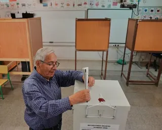 A man wearing glasses places a ballot into a white ballot box at a polling station