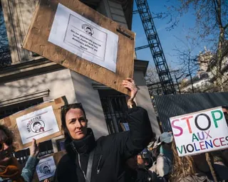 A woman in a black jacket holds up a cardboard sign with a logo and text at a protest