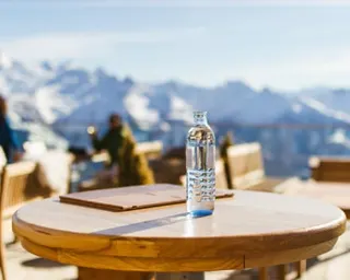 A glass bottle sof water sits on a wooden table with snow-capped mountains in the background