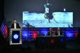 A man at a Nasa podium presents in front of a large screen showing a lunar lander, with two people seated at a table