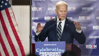 Joe Biden at a podium giving a speech, blue backdrop with BIDEN logos, American flag on the left, wearing a dark suit and striped tie, gesturing with his hands.