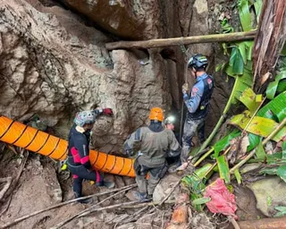Four people in helmets and wetsuits stand in a rocky cave entrance with rescue equipment and vegetation