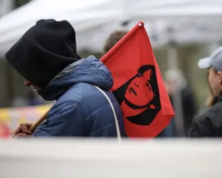 A man wearing a hoodie carrying a red flag bearing an image of Daniela Klette over his shoulder.