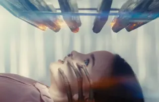 little girl lying on her back in a clinical setting looking up at an array of rods and lights comes down toward her. Spindly alien fingers are touching her face from below.
